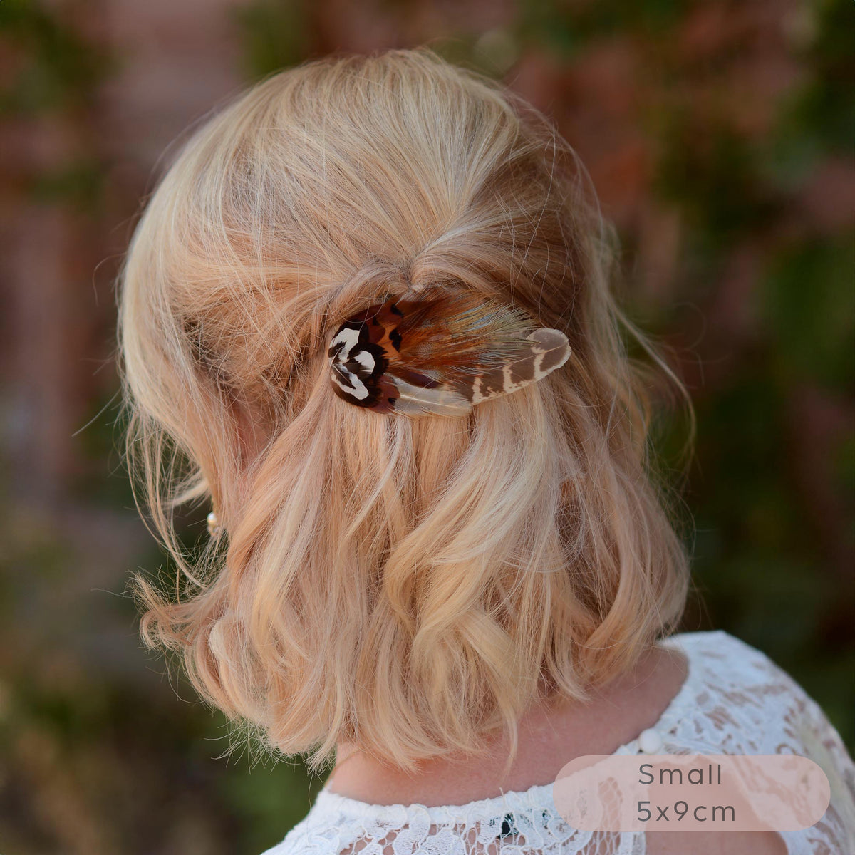A small  wing-shaped pheasant feather hair clip with a black, brown and white patterned feathers, shown securing a blonde woman's half-up hairstyle outdoors. 