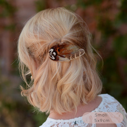 A small  wing-shaped pheasant feather hair clip with a black, brown and white patterned feathers, shown securing a blonde woman's half-up hairstyle outdoors. 