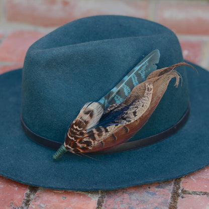 A dark teal felt wide-brim hat with a black band and a large decorative pheasant feather hat pin, resting on a brick surface.
