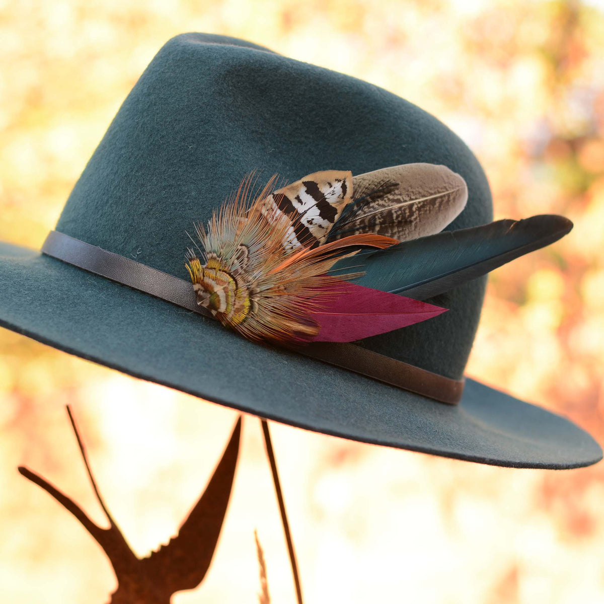 A teal felt wide-brim hat with a black band and decorative feather brooch hat pin in orange, brown, black, teal and burgundy, photographed against a warm blurred outdoor background.