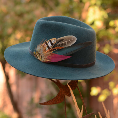 A teal blue felt fedora hat with a black band and decorative hat pin of pheasant feathers in teal, burgundy, brown, and white, displayed outdoors against a blurred natural background.