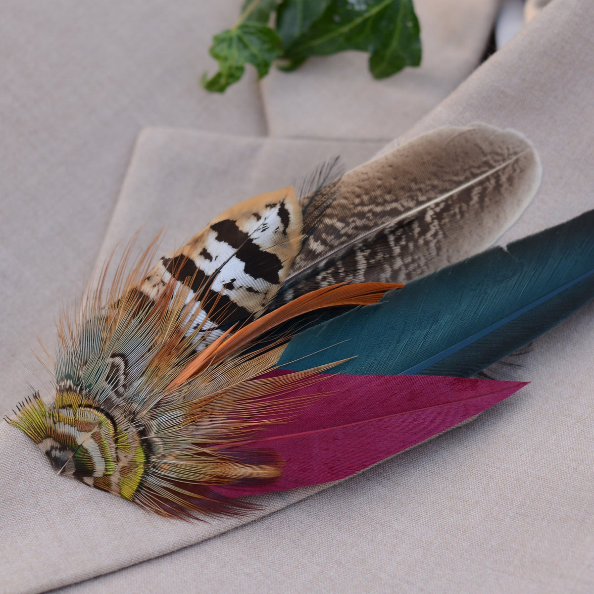 A decorative feather hat pin with multiple colourful feathers, including a burgundy feather, teal-blue feather, brown patterned feather, and tan-and-black striped feather, resting on a beige fabric surface.