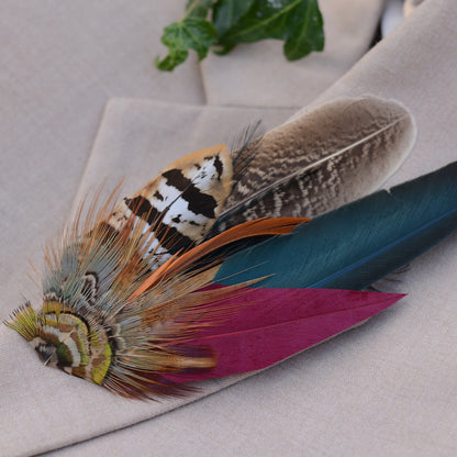A decorative feather hat pin with multiple colourful feathers, including a burgundy feather, teal-blue feather, brown patterned feather, and tan-and-black striped feather, resting on a beige fabric surface.