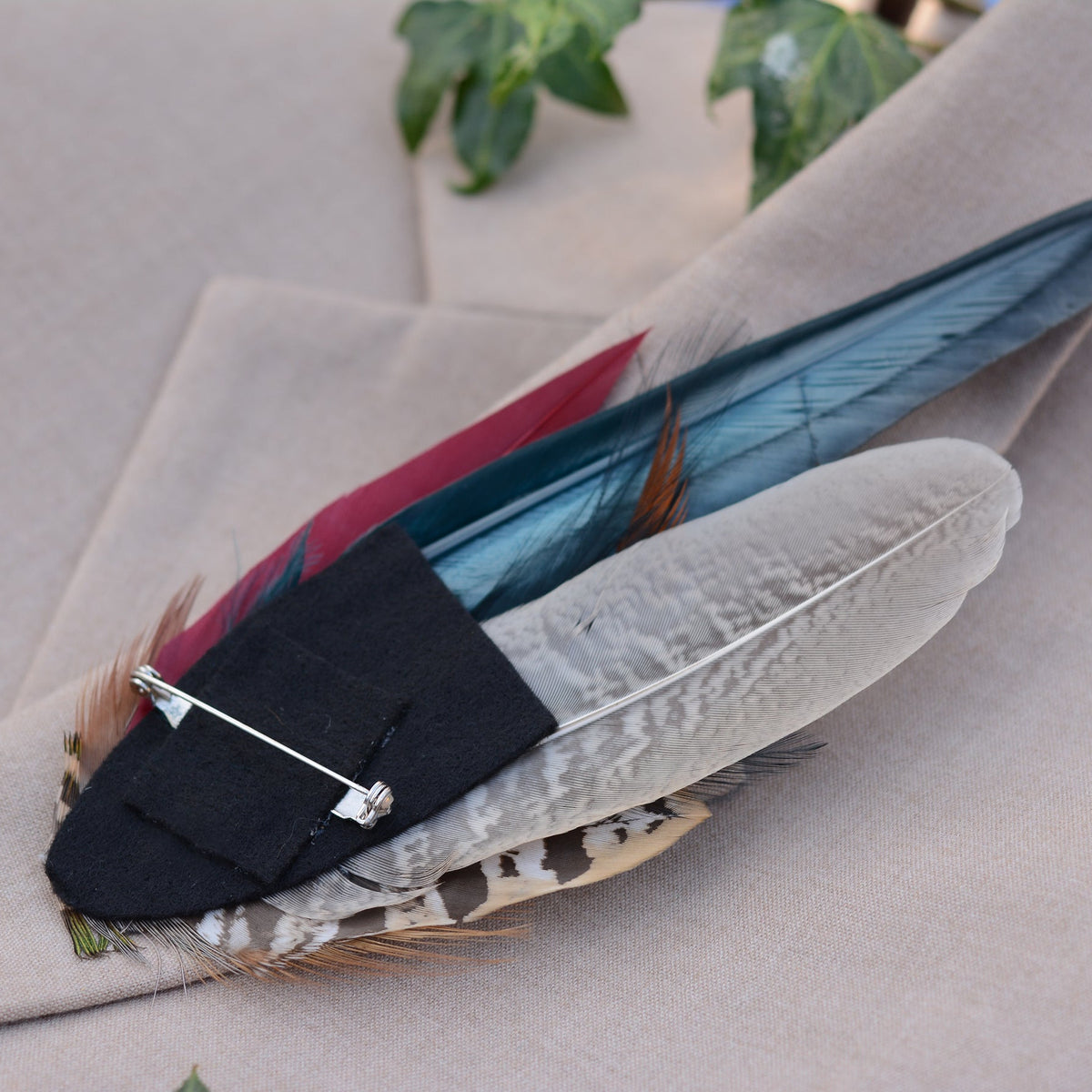 A feathered brooch or hat pin with a black felt base and silver safety pin, displayed on a beige fabric surface.