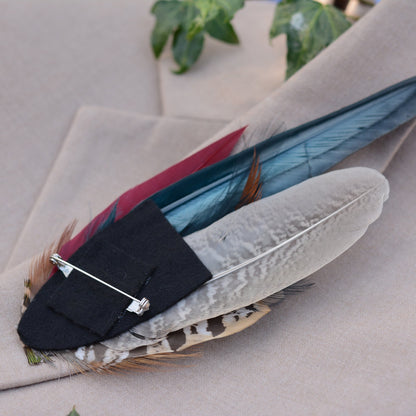 A feathered brooch or hat pin with a black felt base and silver safety pin, displayed on a beige fabric surface.