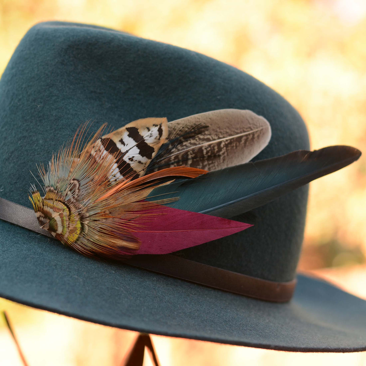 A close-up of a wide-brimmed felt hat in muted teal blue, decorated with a black band and a feathered accent featuring a brown-and-white patterned feathers, teal  and a burgundy feather, set against a warm, blurred outdoor background.