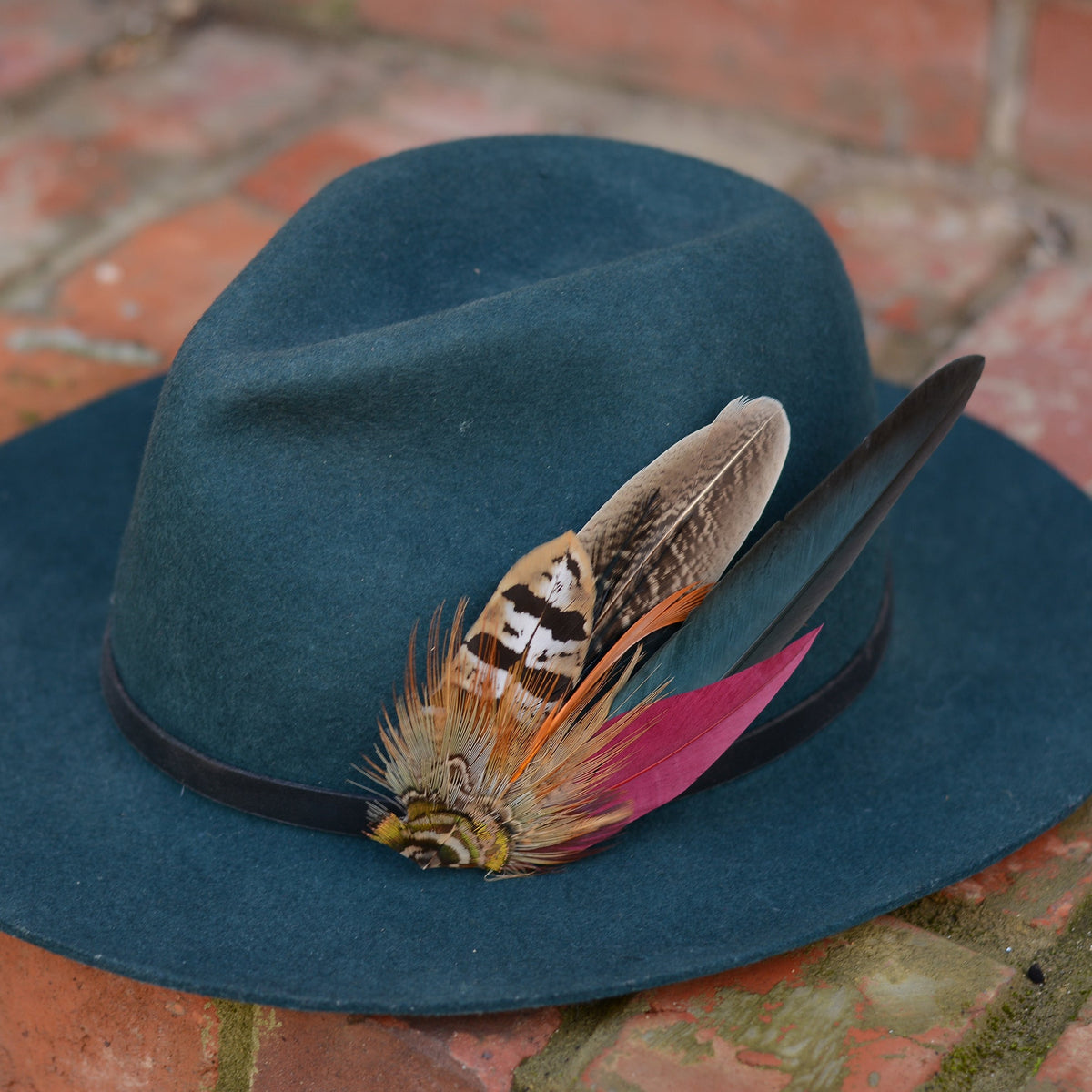 Teal hat with a pheasant feather hat pin on a brick surface