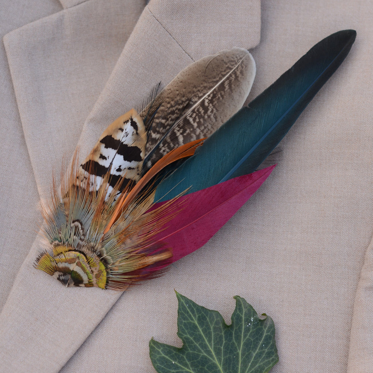 A decorative feather brooch accessory with multiple colourful feathers, including teal, burgundy, brown, orange, and cream, arranged on a beige textured fabric background with a green leaf prop.