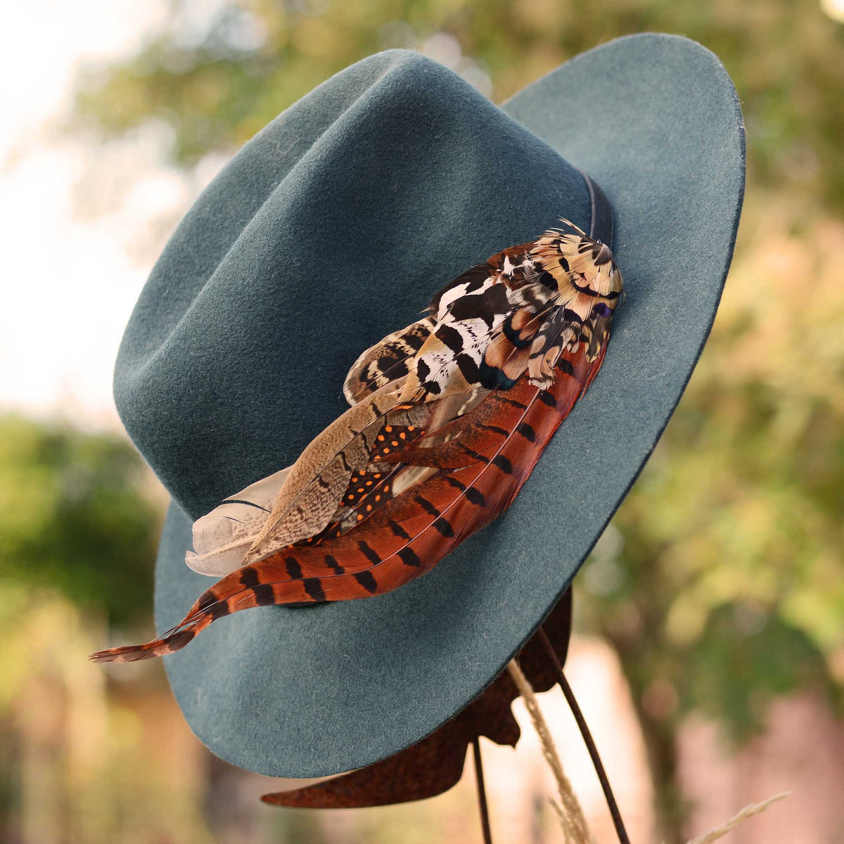 A wide-brimmed boho felt hat in muted blue-green, decorated with a large pheasant feather hat pin and orange-brown feathers, photographed outdoors against a softly blurred natural background.