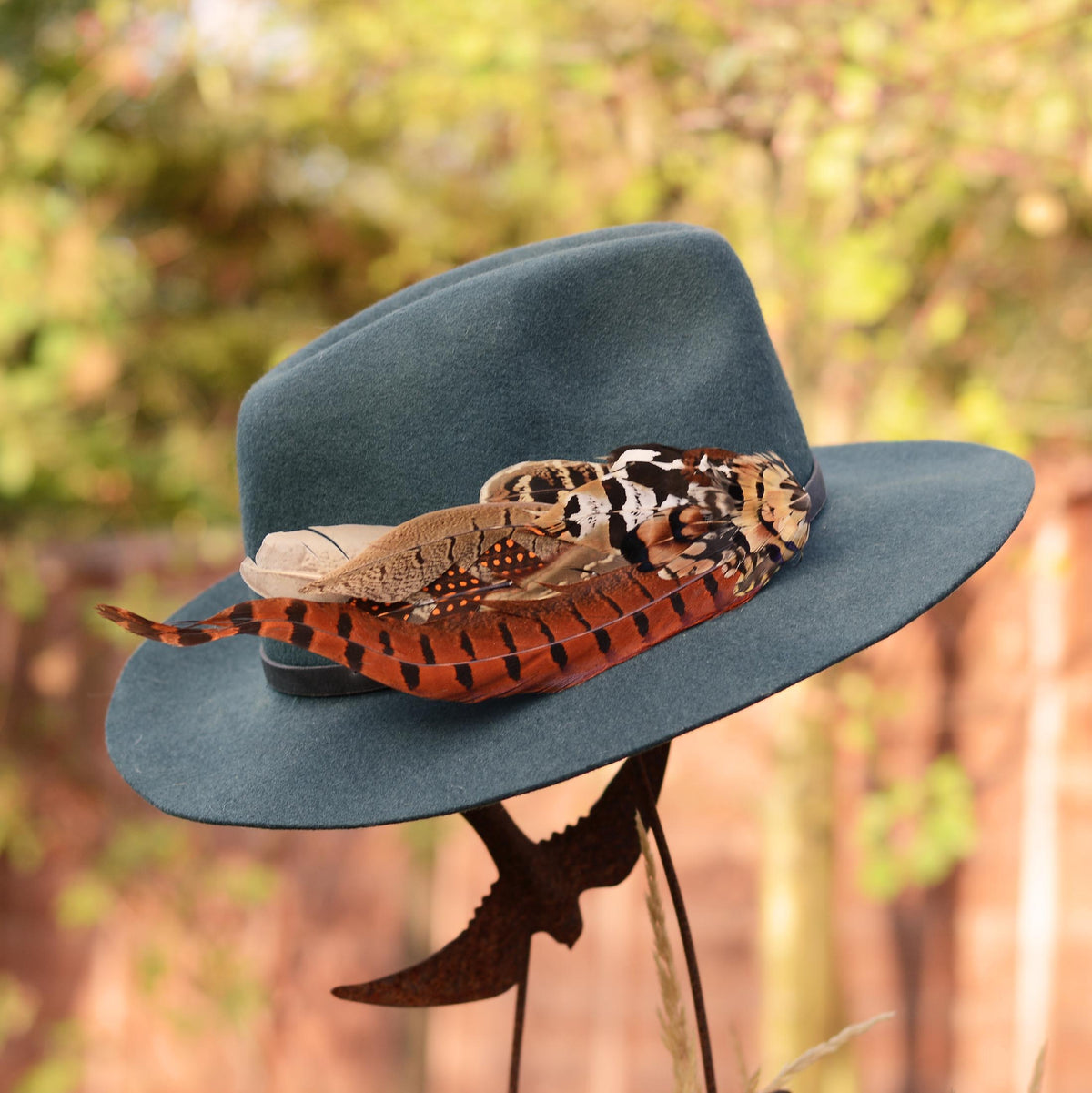 A wide-brimmed felt hat in muted blue-green, decorated with a extra large feather hat decoration in fall colours of rust, orange, black, brown, and cream feathers, photographed outdoors against a blurred natural background.