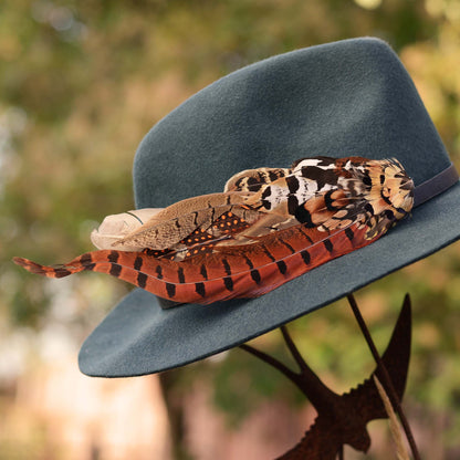 A dark teal felt hat with a black band, decorated with a large pheasant feather hat pin in brown, orange, black, and cream, photographed outdoors against a blurred green and gold background.