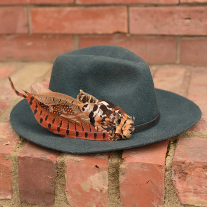 A dark teal felt fedora hat with a black band and a bohemian decorative hat pin of brown, orange, and black patterned feathers, resting on a brick surface against a brick wall.