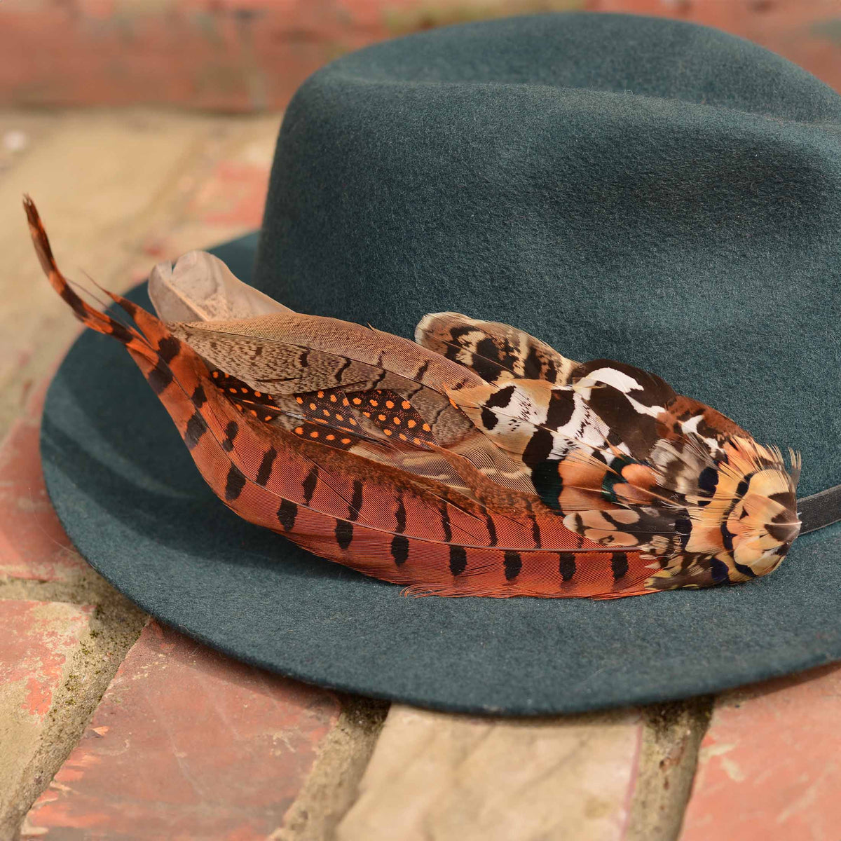 A dark teal felt hat with a large decorative feather hat pin in autumn colours of copper, orange, brown, black, and white, resting on a brick surface.