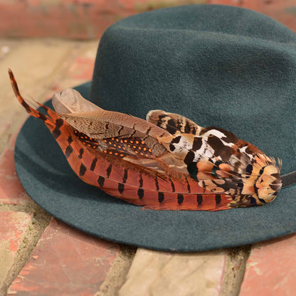 A dark teal felt hat with a large decorative feather hat pin in autumn colours of copper, orange, brown, black, and white, resting on a brick surface.