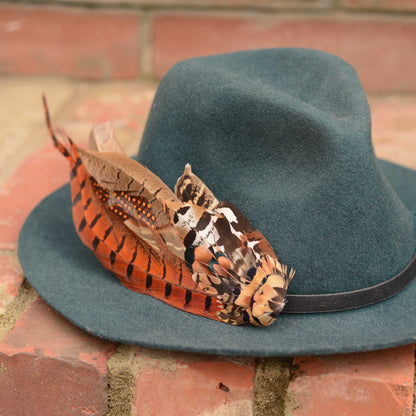 A dark teal felt hat with a black band and  large decorative pheasant feather hat pin mount in rust, burnt orange, brown, black, and white, resting on a red brick surface.