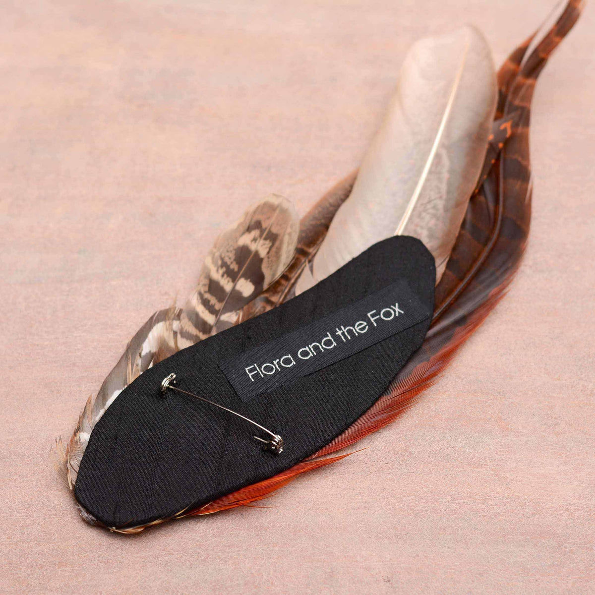 A feather pin by Flora and the Fox, featuring a black slik-covered base with a silver brooch pin with roll lock clasp and a white embroidered brand label.