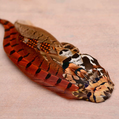 A decorative feather hat pin with a warm burnt orange and rust colour palette and black tiger-like stripes, layered with multicoloured patterned feathers in shades of brown, copper, beige, black, and white, resting on a textured light pink surface.