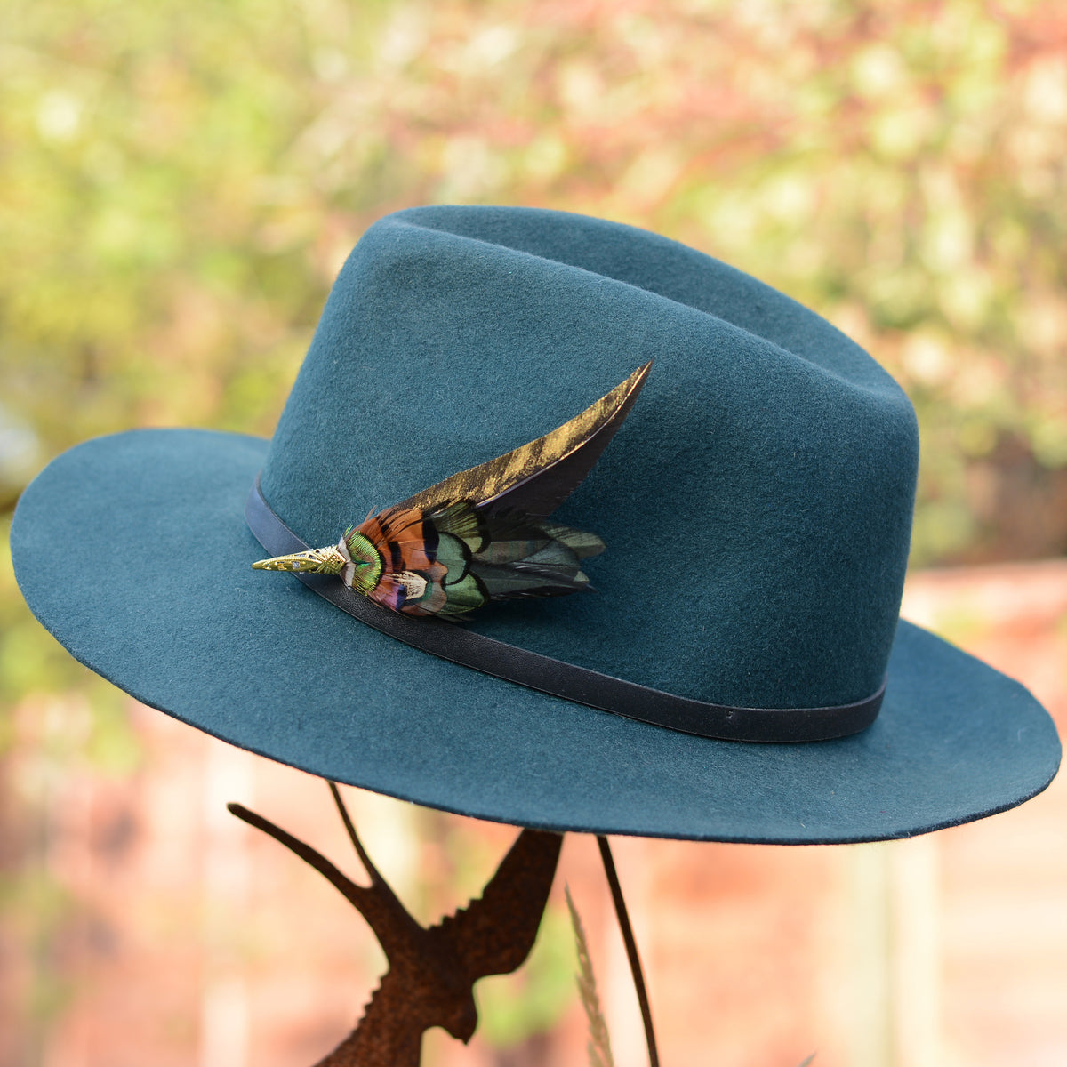 A teal blue felt cowboy hat with a black band and a decorative feather hat pin accent, displayed on a rustic metal stand against a blurred outdoor background.