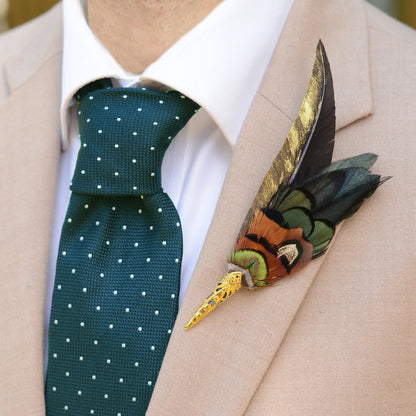 A close-up of a men's formal outfit featuring a dark green tie with white polka dots, a beige suit jacket, and a decorative feathered lapel pin on the chest.