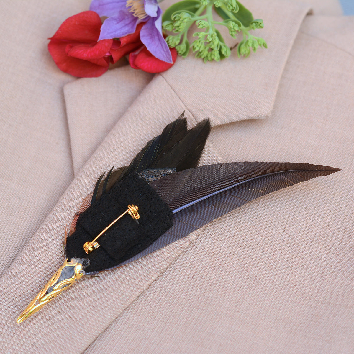 A decorative feather pin with a black felt base, gold-tone metal cap and pin hardware, on beige fabric with red and purple floral accents.
