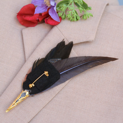A decorative feather pin with a black felt base, gold-tone metal cap and pin hardware, on beige fabric with red and purple floral accents.