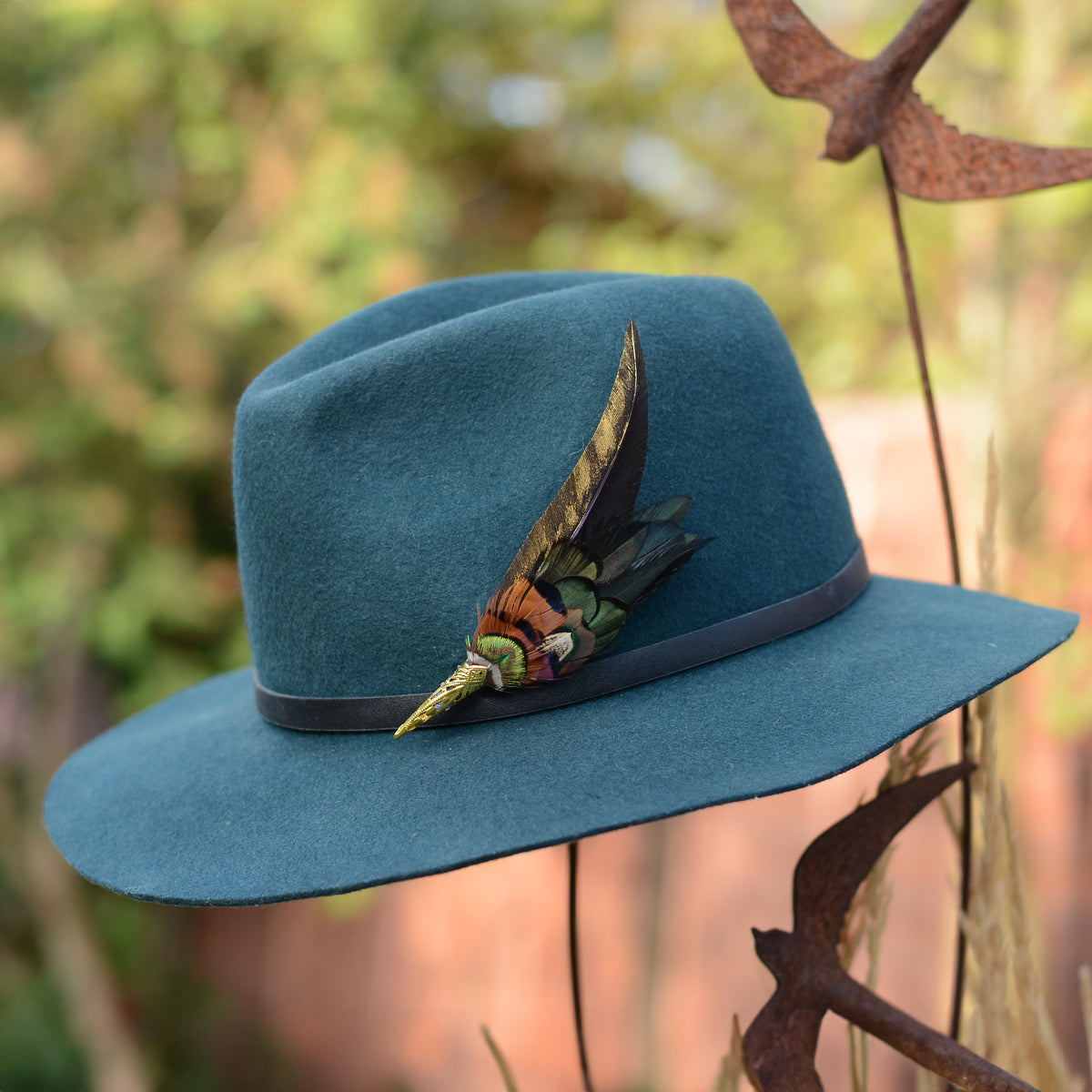 A teal blue felt wide-brim hat with a black band and a decorative feather hat pin photographed outdoors against a blurred natural background.