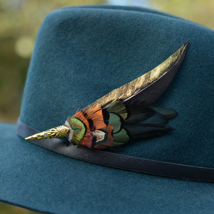 A close-up of a dark teal felt hat with a black band, decorated with a large feather brooch embellishment featuring metallic gold, green, orange, and black feathers.