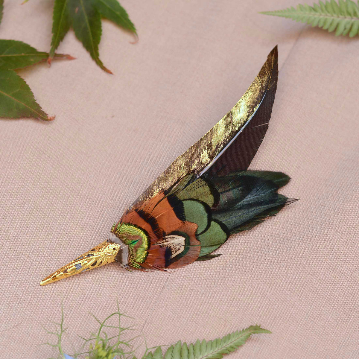 A decorative feather pin with a long gold-painted feather and layered feathers in green, orange, black, and metallic gold, displayed on a beige fabric background with green leaves.
