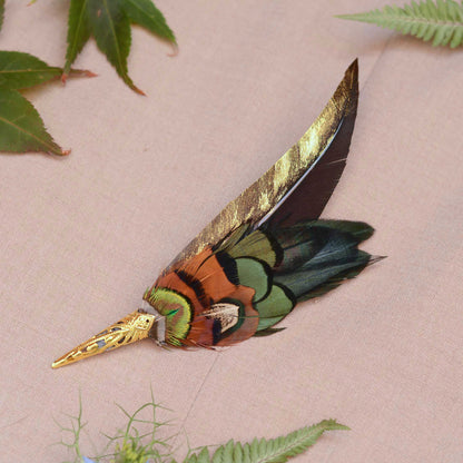 A decorative feather pin with a long gold-painted feather and layered feathers in green, orange, black, and metallic gold, displayed on a beige fabric background with green leaves.