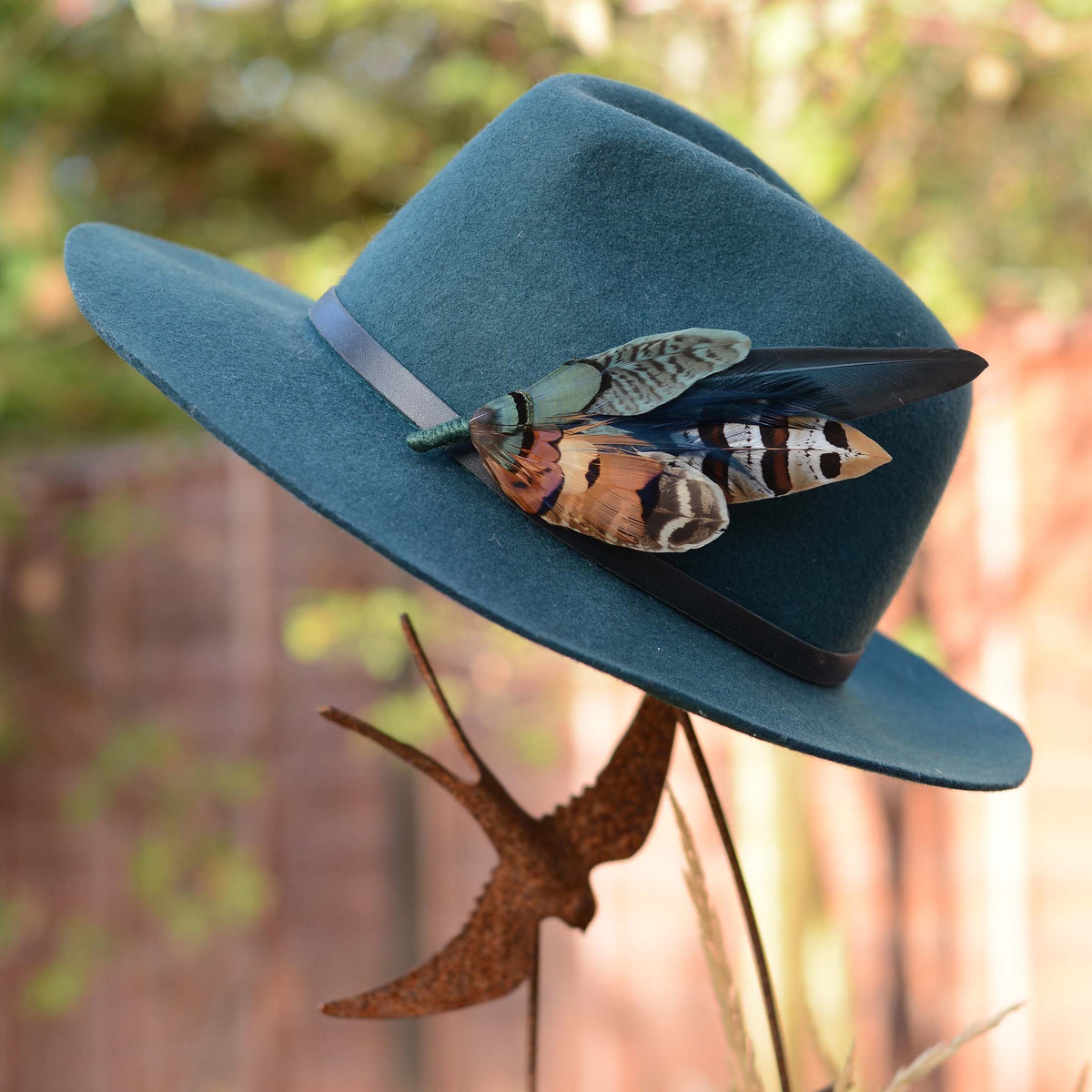 Large decorative pheasant feather hat pin in coppers and greens pinned on a teal wool fedora on displayed on a steel diving swallow stand outside in morning light.