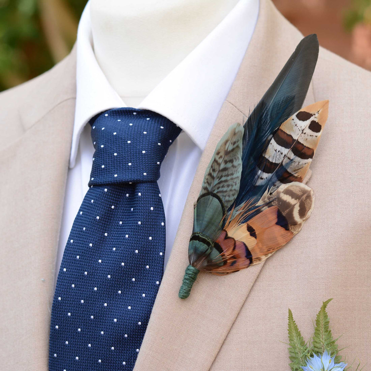 A close-up of a beige suit jacket on a mannequin or model, styled with a white dress shirt, a navy blue polka-dot tie, and a large decorative feathered boutonniere on the lapel.