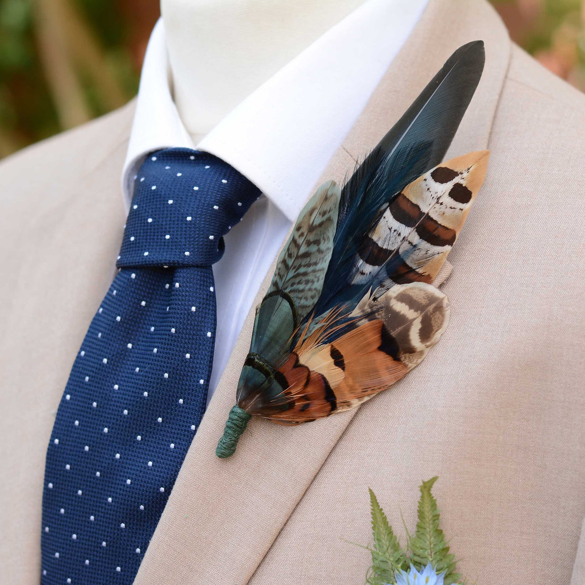 A close-up of a beige suit jacket on a mannequin or torso form, styled with a white dress shirt, a navy polka-dot tie, and a large decorative feather boutonniere pin in green and copper pheasant feathers.
Perfect for autumn / winter racing or a keepsake grooms buttonhole