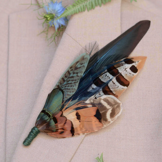 A decorative feather brooch pin in brown, beige, black, green and teal tones, with a smaller iridescent green feather attached, displayed on a light beige fabric background with a small blue flower prop.