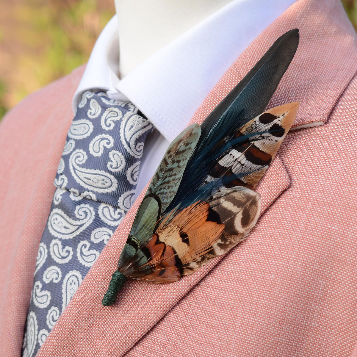 A close-up of a large decorative lapel pin in green and copper pheasant feathers, pinned to a men's suit jacket in pinkish salmon linen, styled with a white collared shirt and a blue paisley tie.