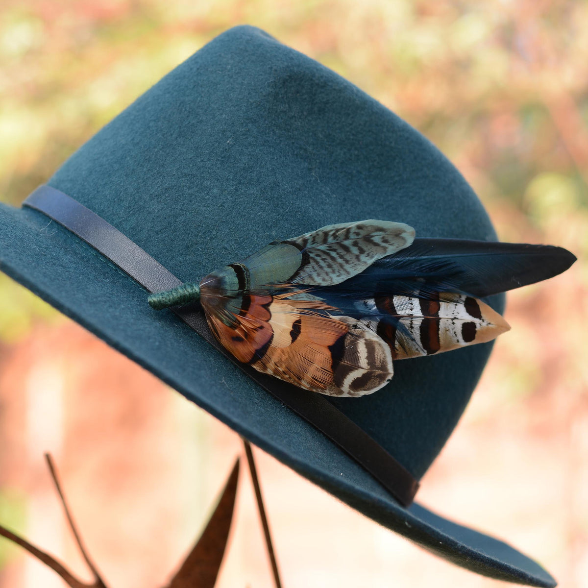 A dark teal felt hat with a black band, decorated with a large, statement feather hat pin, photographed against a warm, blurred outdoor background. A perfect gift for someone who enjoys pheasant hunting.