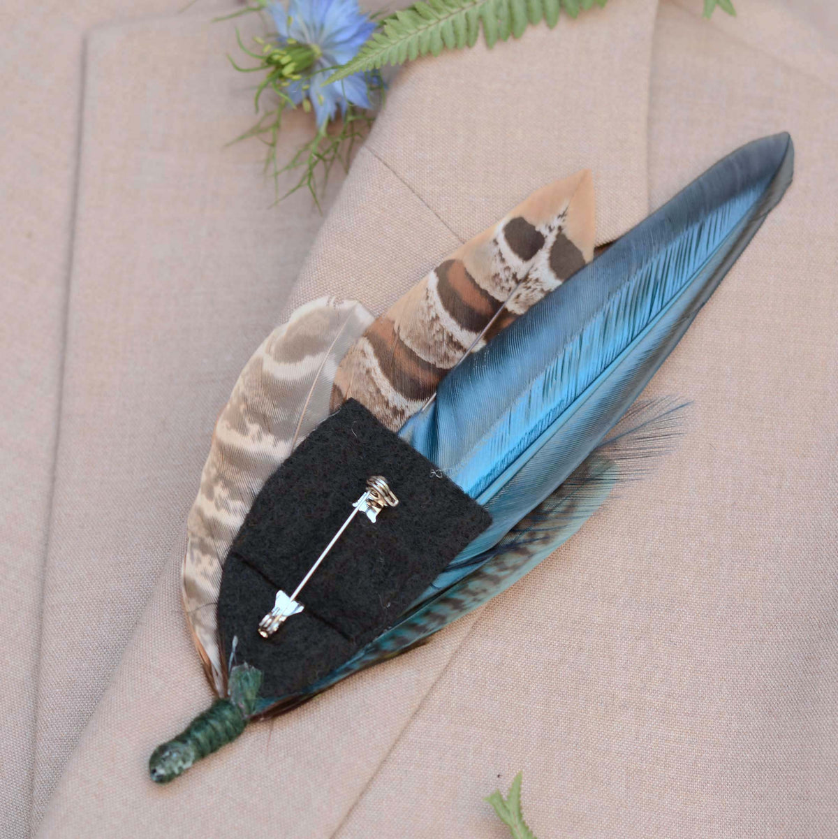 A decorative feather brooch pin with multiple feathers in blue, brown, beige, and green tones, attached to a black felt base with a silver safety pin, displayed on a beige textured fabric background.