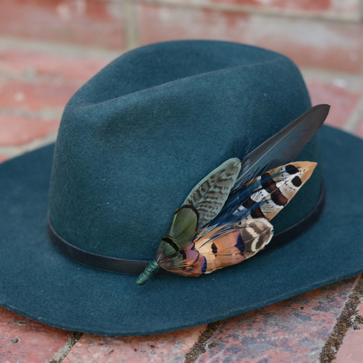A dark teal felt fedora hat with a black band, decorated with a large statement pheasant feather hat pin, resting on a red brick surface.