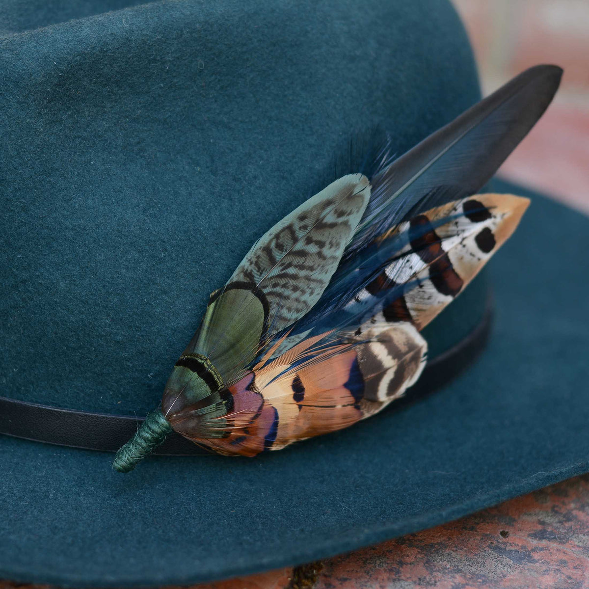 A dark teal wide-brim hat with a black band, decorated with a statement feather hat pin: one green and brown patterned, and one blue, black, and orange-and-white spotted.