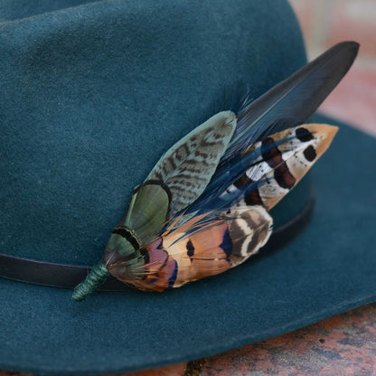 A dark teal wide-brim hat with a black band, decorated with a statement feather hat pin: one green and brown patterned, and one blue, black, and orange-and-white spotted.