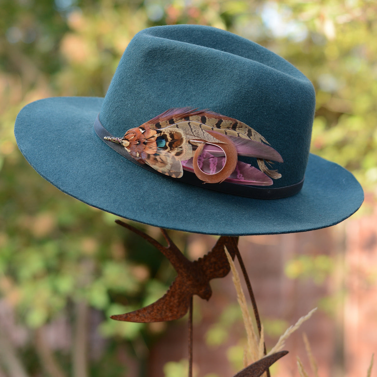 A statement purple and pheasant feather hat pin pinned on a teal blue felt wide-brim hat photographed outdoors against a blurred green and brown garden background.