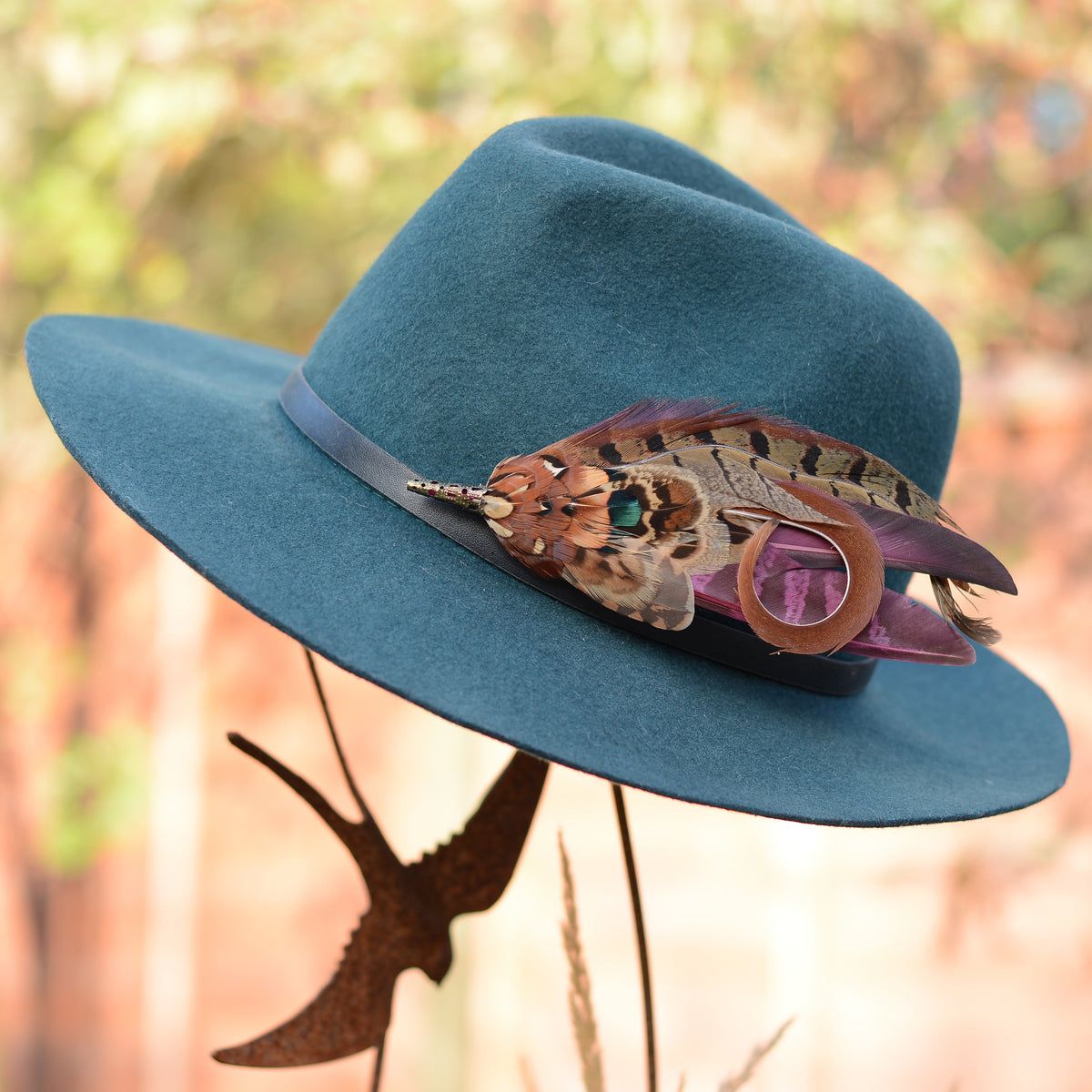 A teal blue felt wide-brim hat with a dark band and an ornate pheasant feather hat pin embellishment, displayed against a blurred outdoor background.