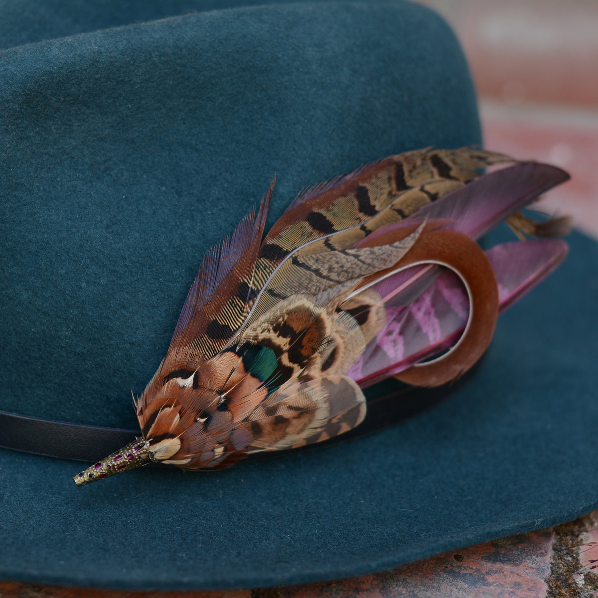 A dark teal felt hat with a black band, decorated with a large brown and tan pheasant feather brooch embellishment featuring black stripes and a small iridescent green accent.