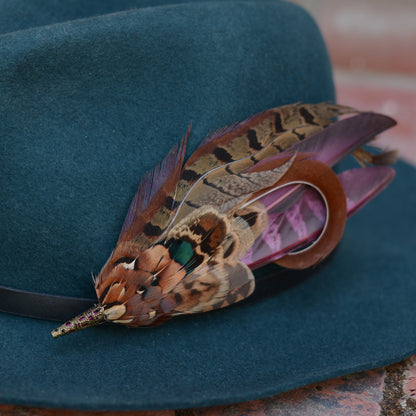 A dark teal felt hat with a black band, decorated with a large brown and tan pheasant feather brooch embellishment featuring black stripes and a small iridescent green accent.