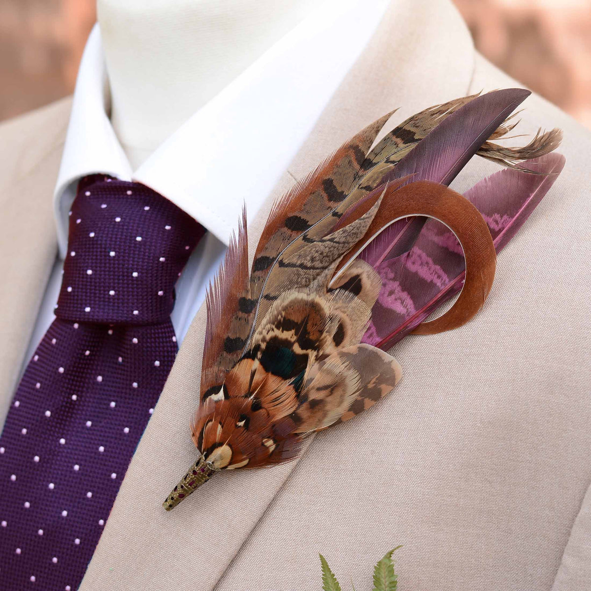 A decorative feathered lapel pin with brown, black, and purple pheasant feathers, pinned to a beige suit jacket over a white dress shirt and a purple polka-dot tie.