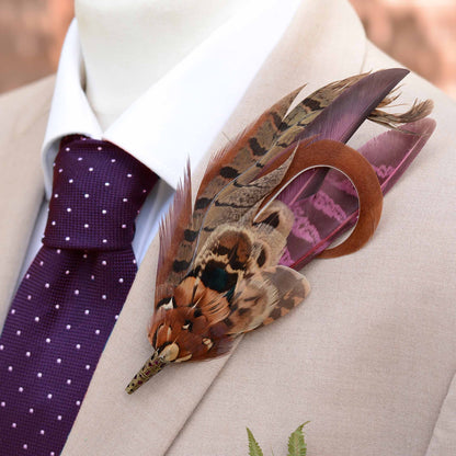 A decorative feathered lapel pin with brown, black, and purple pheasant feathers, pinned to a beige suit jacket over a white dress shirt and a purple polka-dot tie.