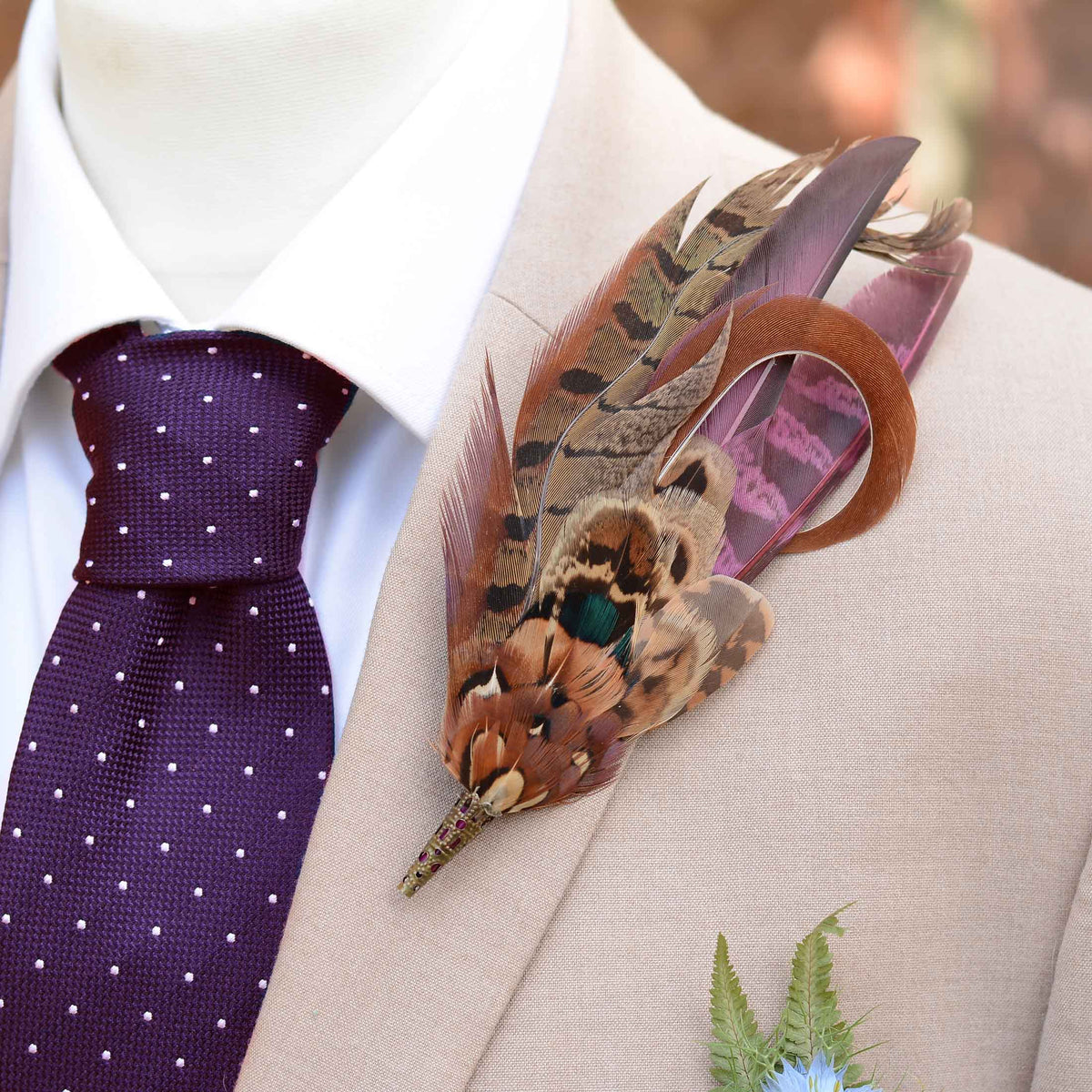A close-up of a men's formal outfit featuring a beige suit jacket, a white dress shirt, a purple polka-dot tie, and a decorative feathered boutonniere pin on the lapel.