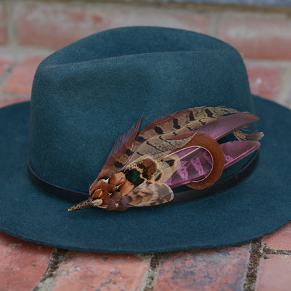 A dark teal felt hat with a black band, decorated with a large brown an purple pheasant feather hat pin, resting on a red brick surface.