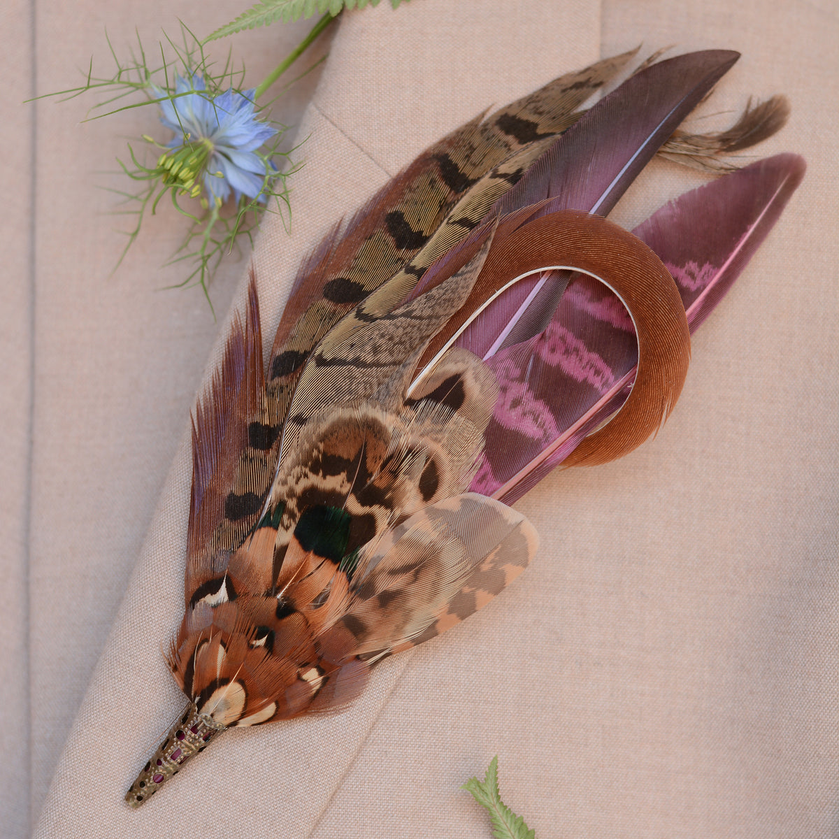 A decorative feather accessory with layered brown, tan, and burgundy patterned pheasant feathers displayed on a beige fabric background with a small blue flower prop.