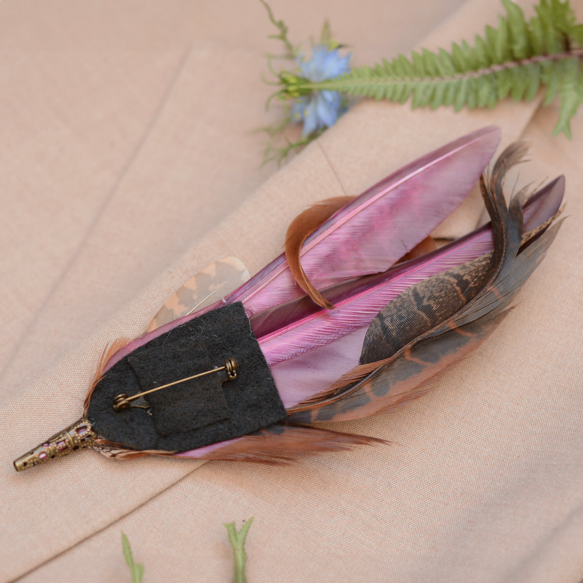 A decorative feather pin featuring purple and brown feathers with a black felt backing and brass-tone safety brooch pin, displayed on a beige textured surface with a fern and small blue flower prop.
