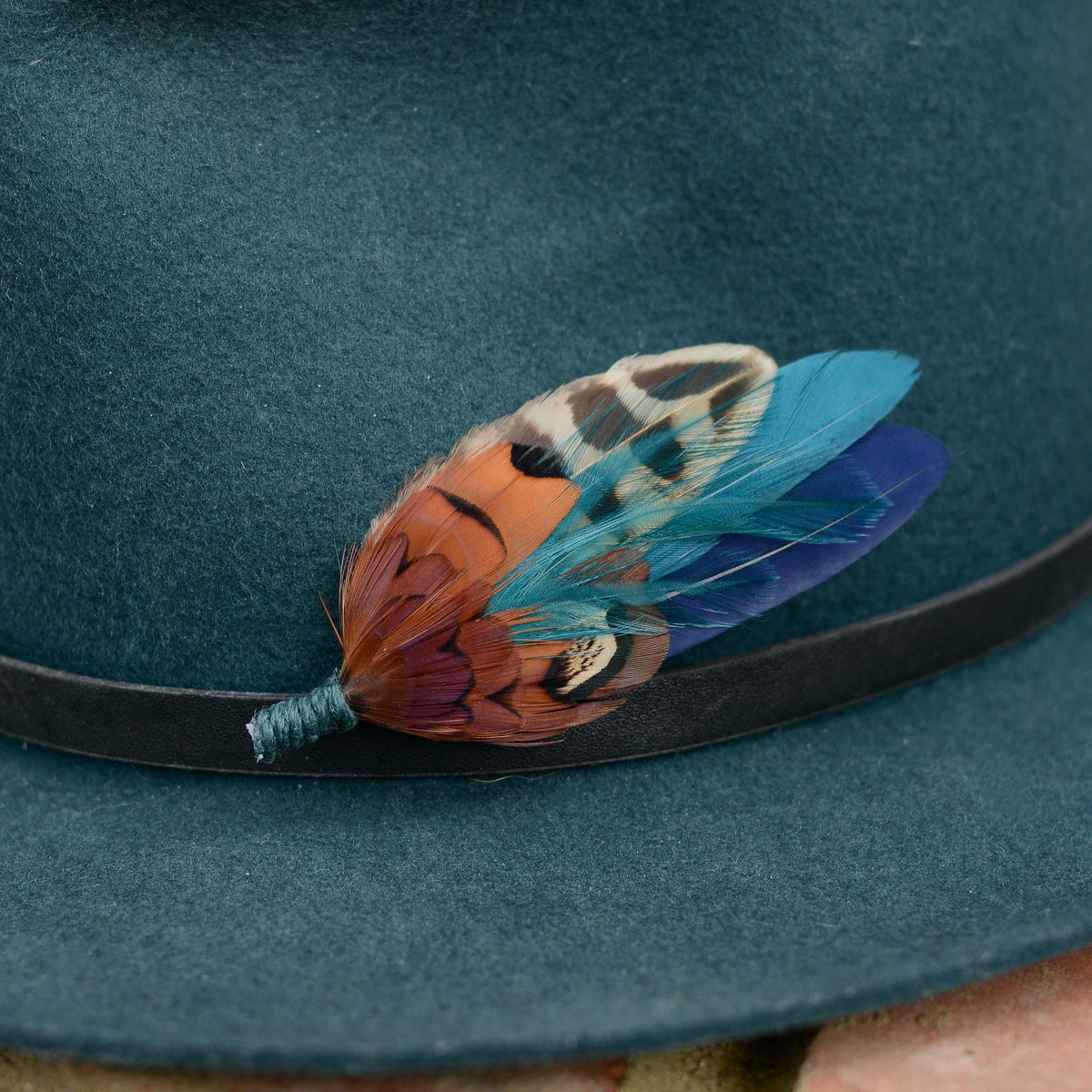 A close-up of a dark teal felt hat with a black band, decorated with a colourful feather pin featuring orange, brown and black patterned pheasant feathers paired with teal and blue.